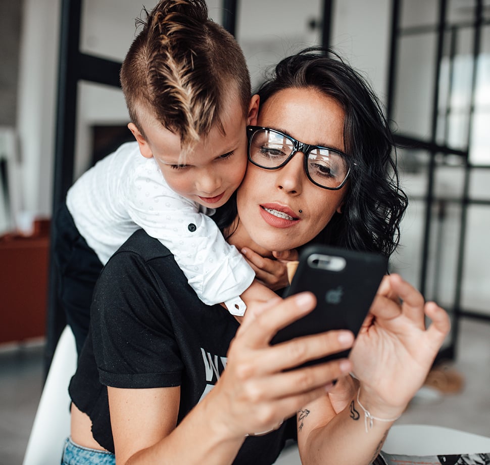 Mutter mit Smartphone und Kind auf dem Rücken organisiert Nachrichten und Familienalltag – praktische Muttertags-Geschenke 2026 für immer erreichbare Mamas.