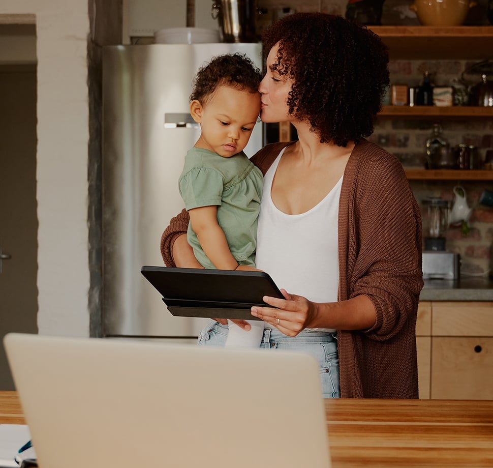 Mutter nutzt ein Tablet in der Küche mit ihrem Kind auf dem Arm – smarte Muttertagsüberraschung und praktische Muttertagsgeschenke für Familien.
