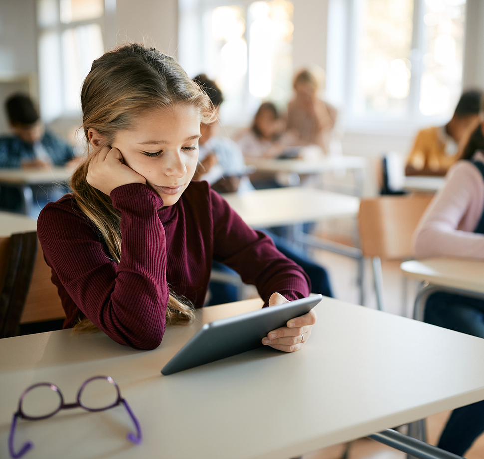 Ein Kind sitzt in einer Schulklasse. Ein Tablet in der Hand.